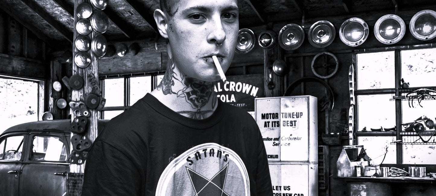 A young gearhead with neck tattoos is in an old auto garage with many old signs, hub caps, and an old car. He is wearing the Satan's All Star T-Shirt from The Asphalt Anarchist Clothing Co. Hot Rod Kustom Kulture Apparel & Products
