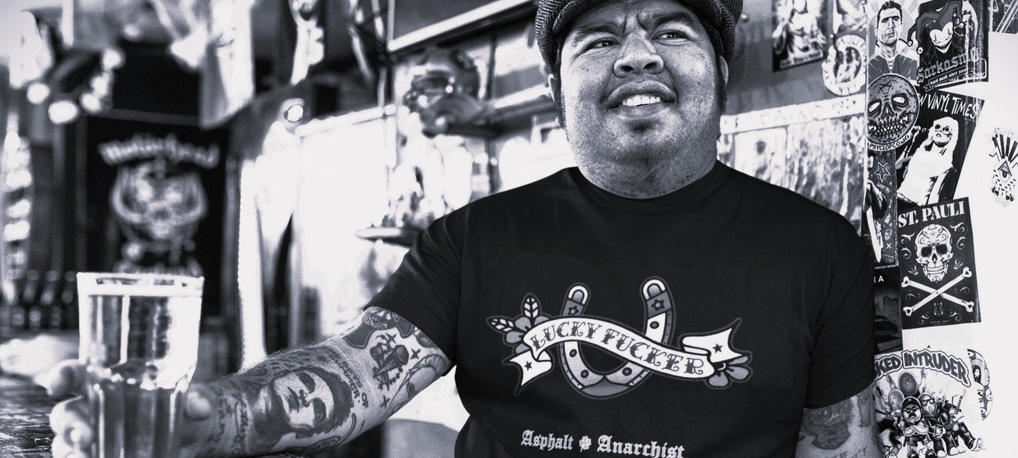 A heavy set Hispanic rockabilly man with tattoos sits at a cool bar enjoying a beer. He is wearing The Lucky Fucker T-Shirt from The Asphalt Anarchist Clothing Co. HOT ROD KUSTOM KULTURE APPAREL & PRODUCTS
